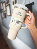 Beige Stanley mug with handle held by a hand against a blurred indoor background